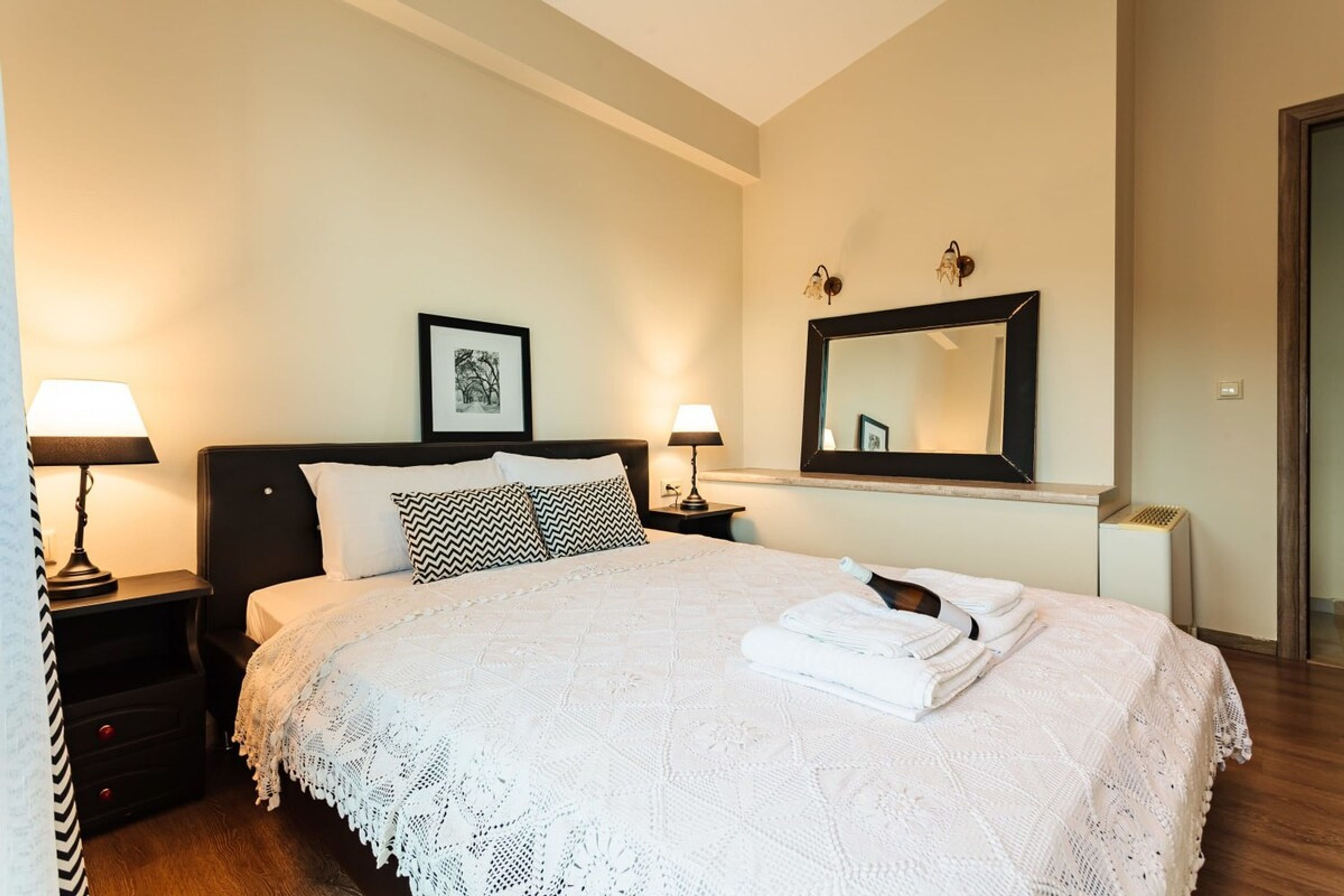 A neatly made bed with a white bedspread and pillows, two bedside lamps, a framed picture on the wall, and a mirror above the bed.