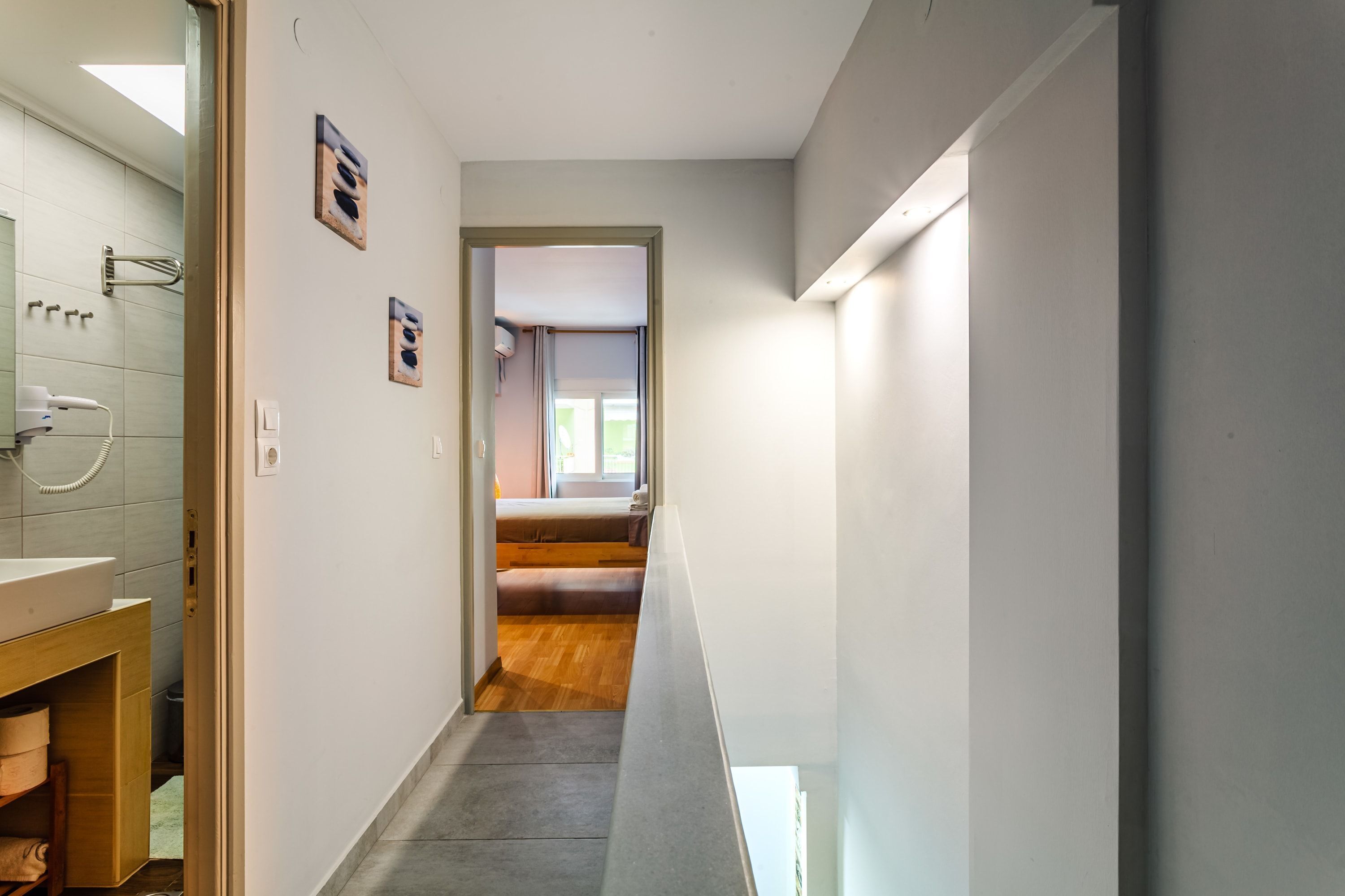 A narrow hallway with a wooden floor, a white wall, and a door leading to a bedroom with a window.