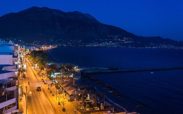 Eine Küstenstadtlandschaft bei Nacht mit einem Berg im Hintergrund, einem Strand und einer beleuchteten Straße mit Gebäuden und Geschäften.