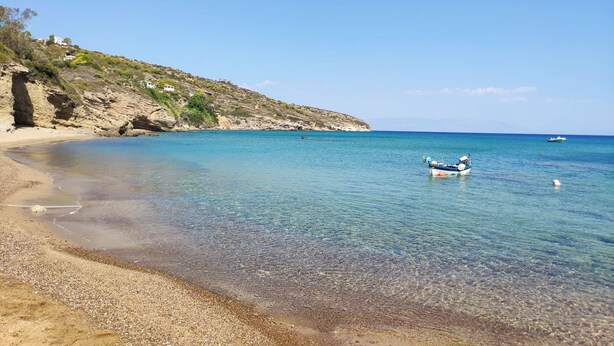 A clear coastal beach with a boat anchored near the shore, rocky cliffs, and a clear blue sky.
