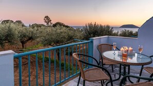 A terrace with wicker chairs and a table, overlooking a vineyard and the sea at sunset.