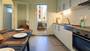 Een kleine keuken met witte kastjes, een inbouwoven, en een eethoek met een houten tafel en stoelen.