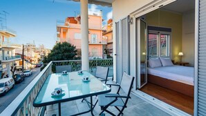 Een balkon met een glazen tafel en stoelen, een bed met uitzicht, en een stadsgezicht op de achtergrond.