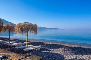 Ein Strand mit Liegestühlen und Reetsonnen schirmchen, ein Kiesstrand und ein ruhiges Meer unter blauem Himmel.