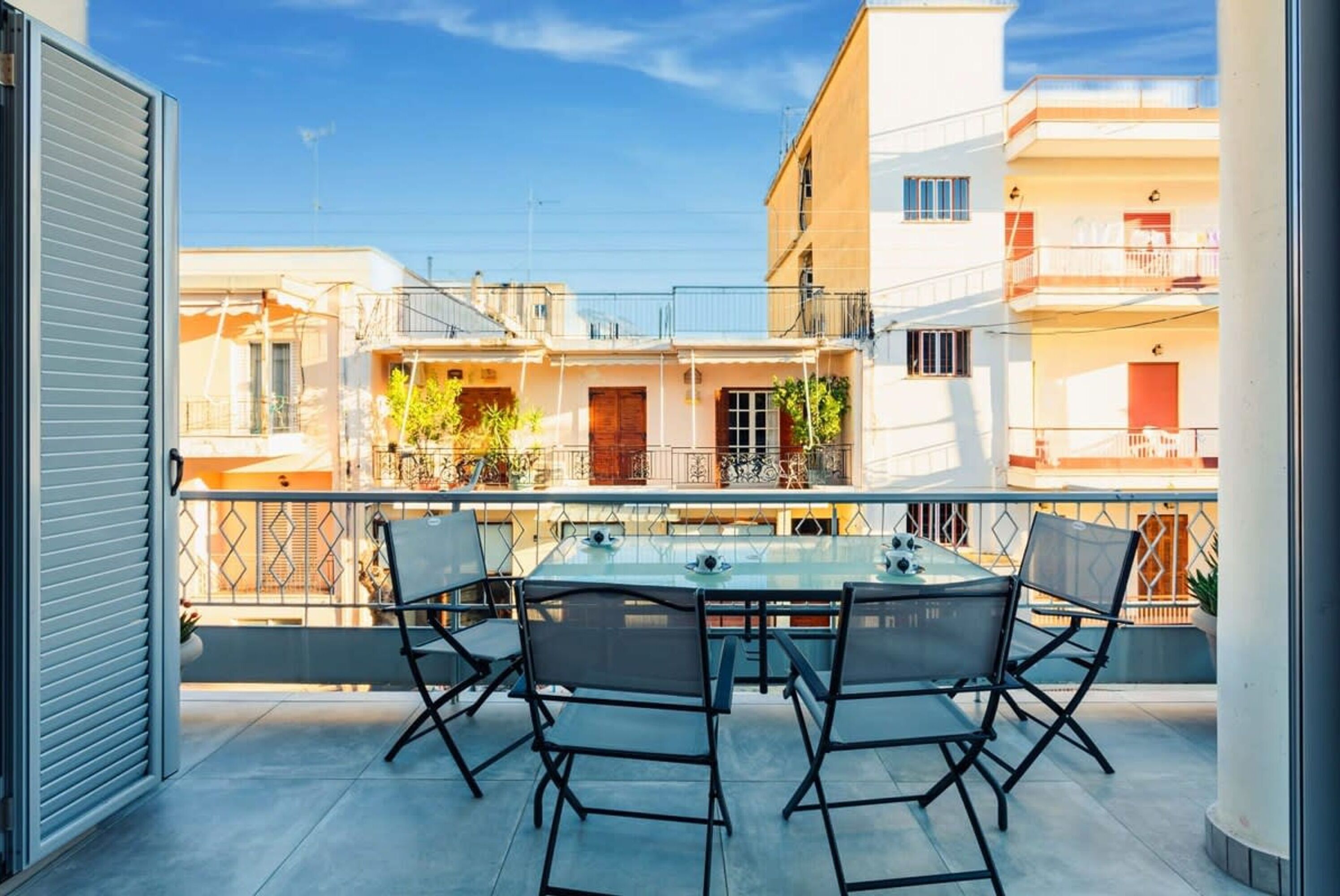 Un balcone con un tavolo e sedie in vetro, affacciato su una zona residenziale con edifici e cielo sereno.