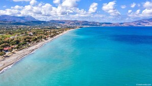 A coastal town with a long beachfront, clear blue sea, and mountains in the background. - Athina Epavlis - Ethereal Charm of Blue Horizons (Platanias)