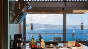 A kitchen with a view of the sea, a coffee maker, a bottle of olive oil, a knife block, and a cheese board. - Athina Epavlis - Ethereal Charm of Blue Horizons (Platanias)