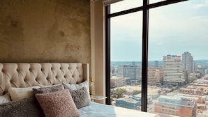 A bedroom with a large bed, a tufted headboard, and a view of a cityscape through large windows.