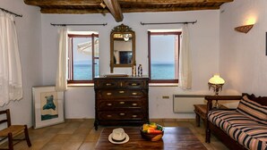 A room with a wooden ceiling, a large mirror, a dresser, a sofa, a table with a hat and fruit bowl, and a view of the sea through two windows. - Waveside Sanctuary - Luxurious Seastone Villa (West Mani)