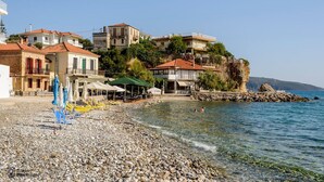 A pebble beach with buildings on a hillside, clear water, and umbrellas. - Waveside Sanctuary - Luxurious Seastone Villa (West Mani)