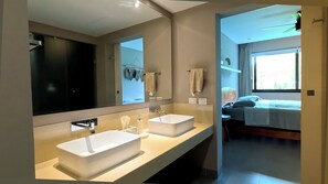 A modern bathroom with two sinks, a large mirror, and a view into a bedroom with a bed and window.