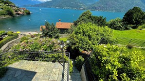 Vista panorámica de un lago con montañas al fondo, un pequeño edificio de techo rojo y un jardín bien cuidado con una escalera de piedra.