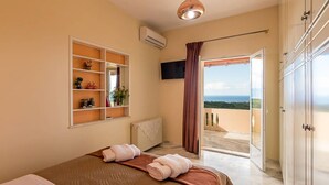 A bedroom with a bed, a shelf with decorative items, an air conditioning unit, a television, and a balcony with a view. - Sea Breeze Stunning Views - Pool Dream Gem (Platanias)