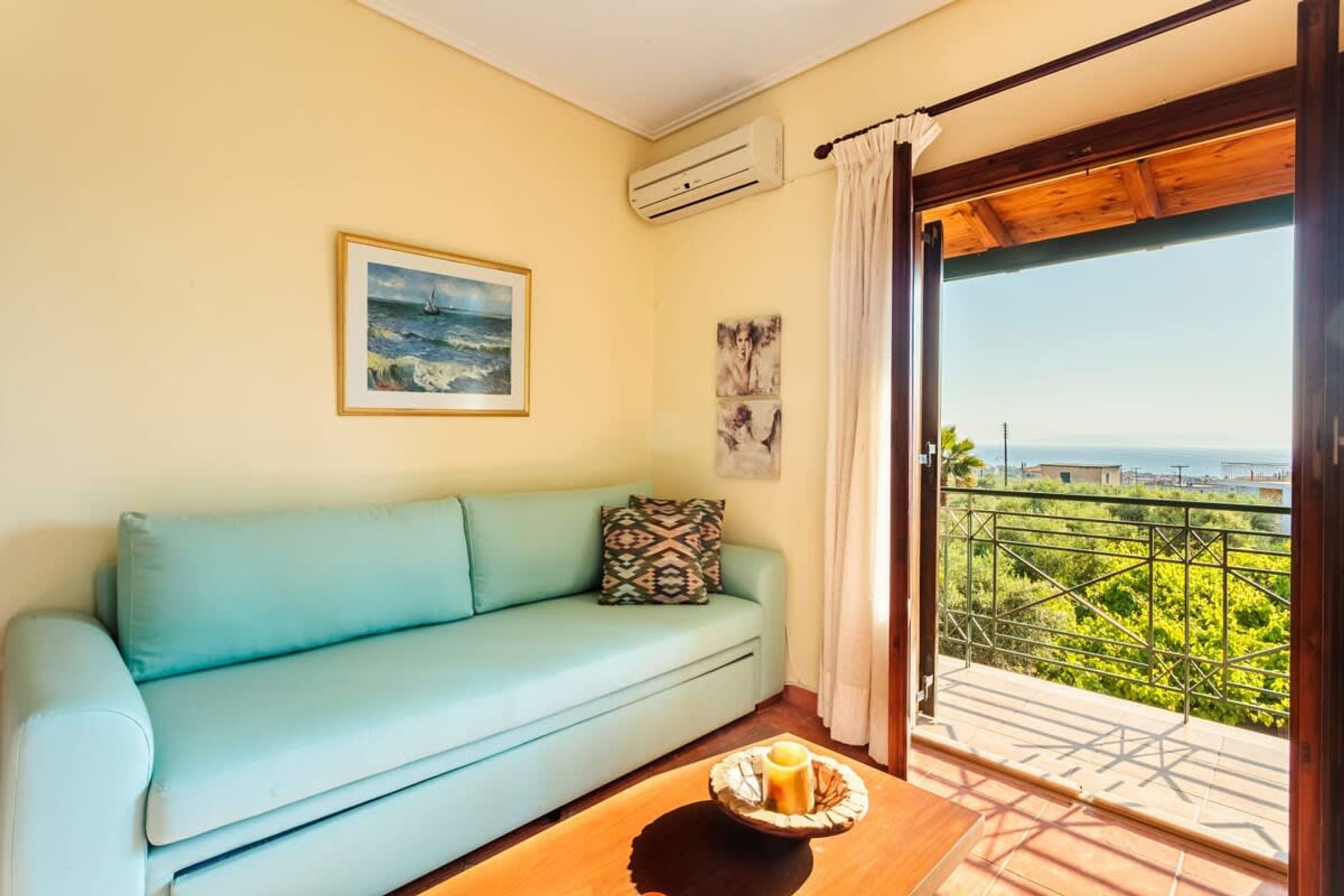 A room with a light blue sofa, a painting of a seascape, and a balcony with a view of greenery and buildings.