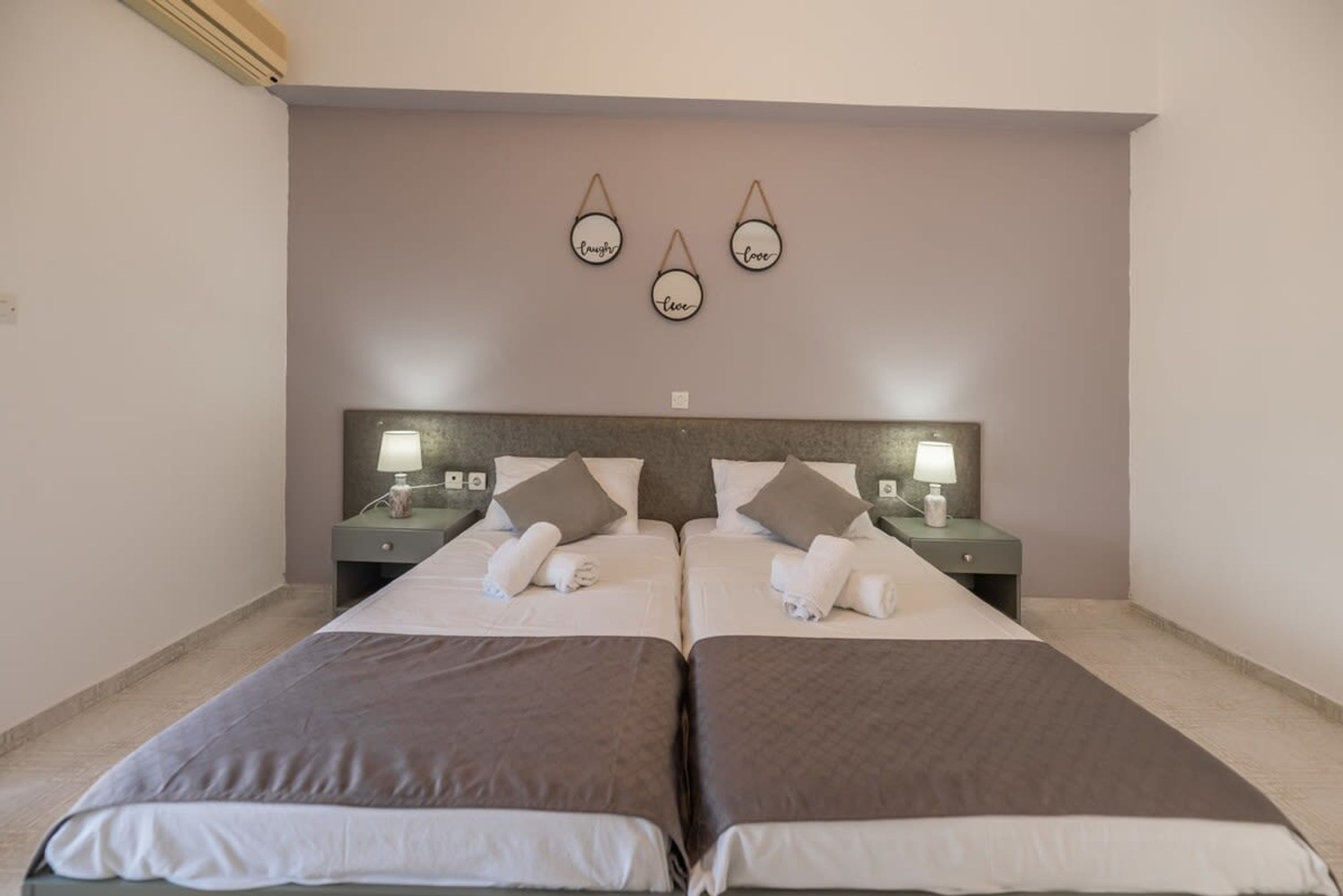 Une chambre double avec deux lits, chacun équipé d’un oreiller et d’une serviette enroulée. Il y a des tables de chevet avec des lampes et un luminaire fixé au mur au-dessus des lits.
