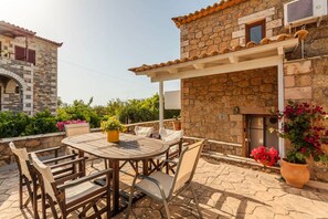 A stone building with a tiled roof, a covered patio with a round table and chairs, and potted plants. - Mani s Aura Seaside-enchanting Stone Gem (West Mani)