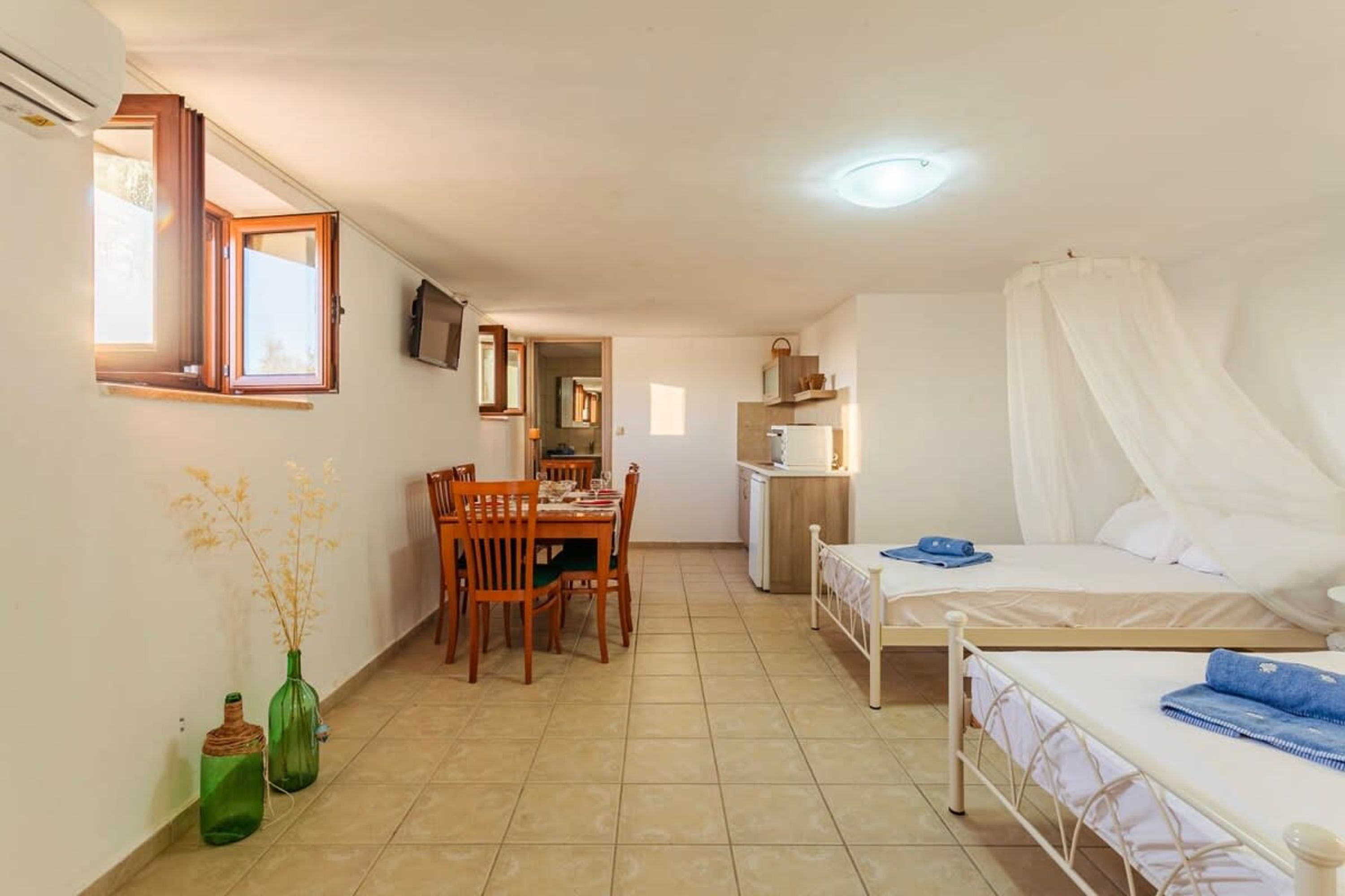 Una habitación pequeña y bien iluminada con cama, una mesa de comedor con sillas, una zona de cocina y un televisor en la pared.