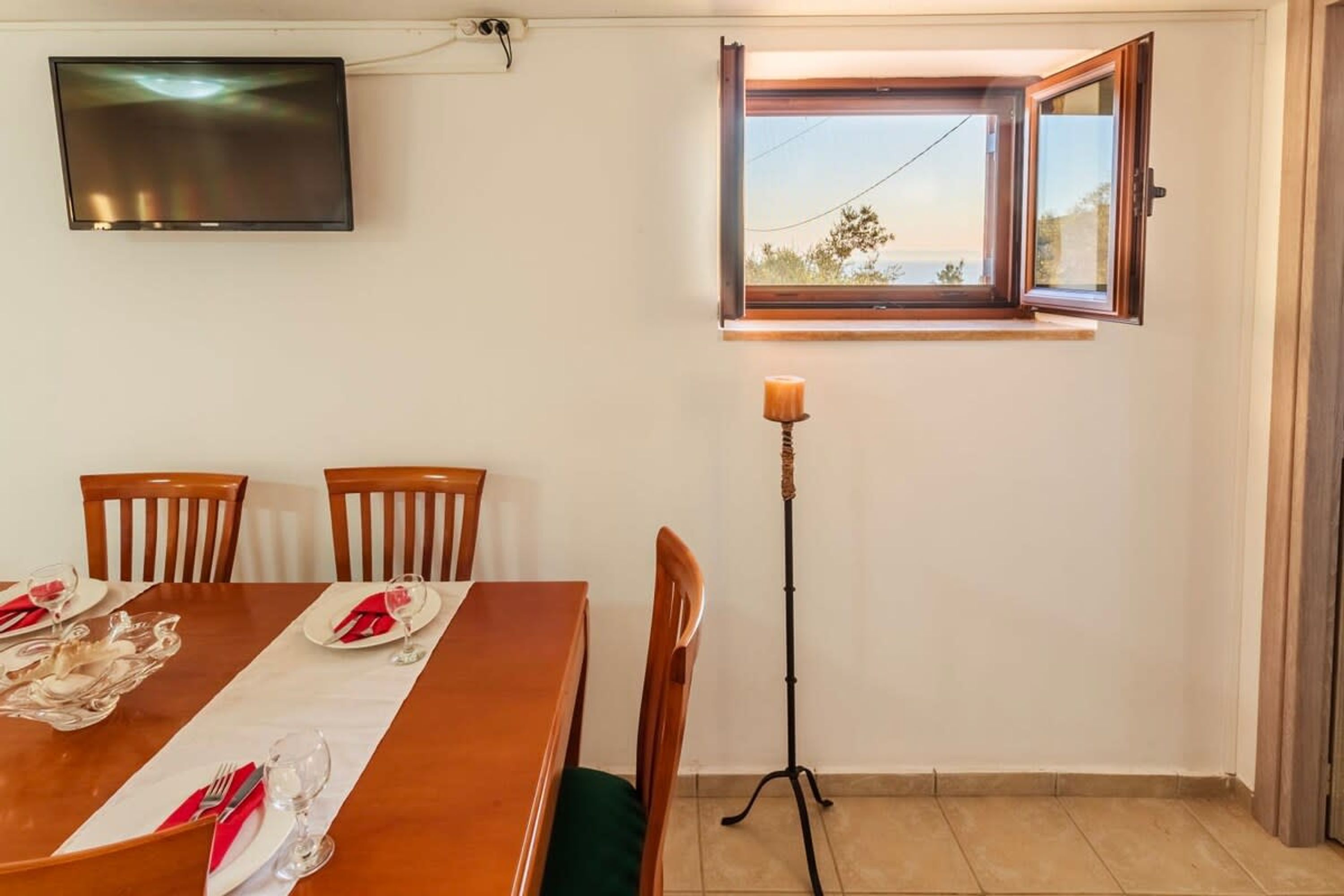Un coin repas avec une table en bois dressée pour quatre personnes, un téléviseur fixé au mur et une fenêtre dont le volet est ouvert.