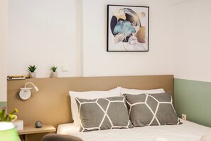 A neatly arranged bedroom with a bed, pillows, a bedside table, a wall-mounted lamp, and a framed artwork.