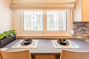 A dining area with a table set for two, a window with a view of buildings, and a potted plant on the left. - Kalamata Lime Studio - Urban Cozy Nest (Kalamata)