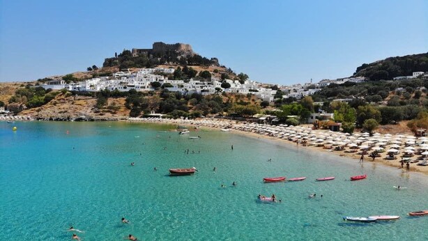 A coastal town with a historic castle on a hill, clear turquoise waters, and a beach with umbrellas and boats.