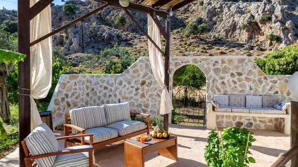 A covered outdoor seating area with a stone wall, a wooden bench, and a table with fruits and a drink. - Atlantic Cave Haven-seaside Fully Equipped (Rhodes)