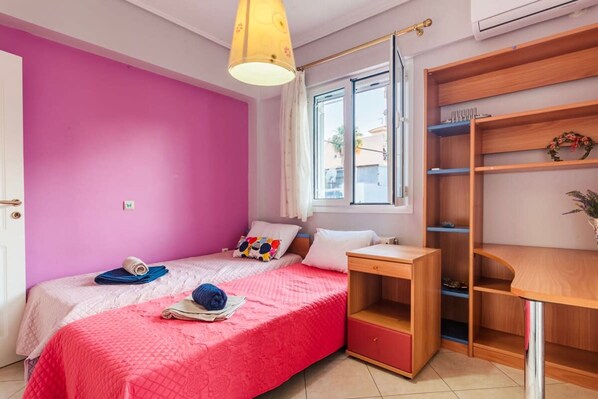 Une chambre avec un mur rose, un lit recouvert d’une couverture rouge, un bureau en bois et une fenêtre donnant sur des bâtiments à l’extérieur.