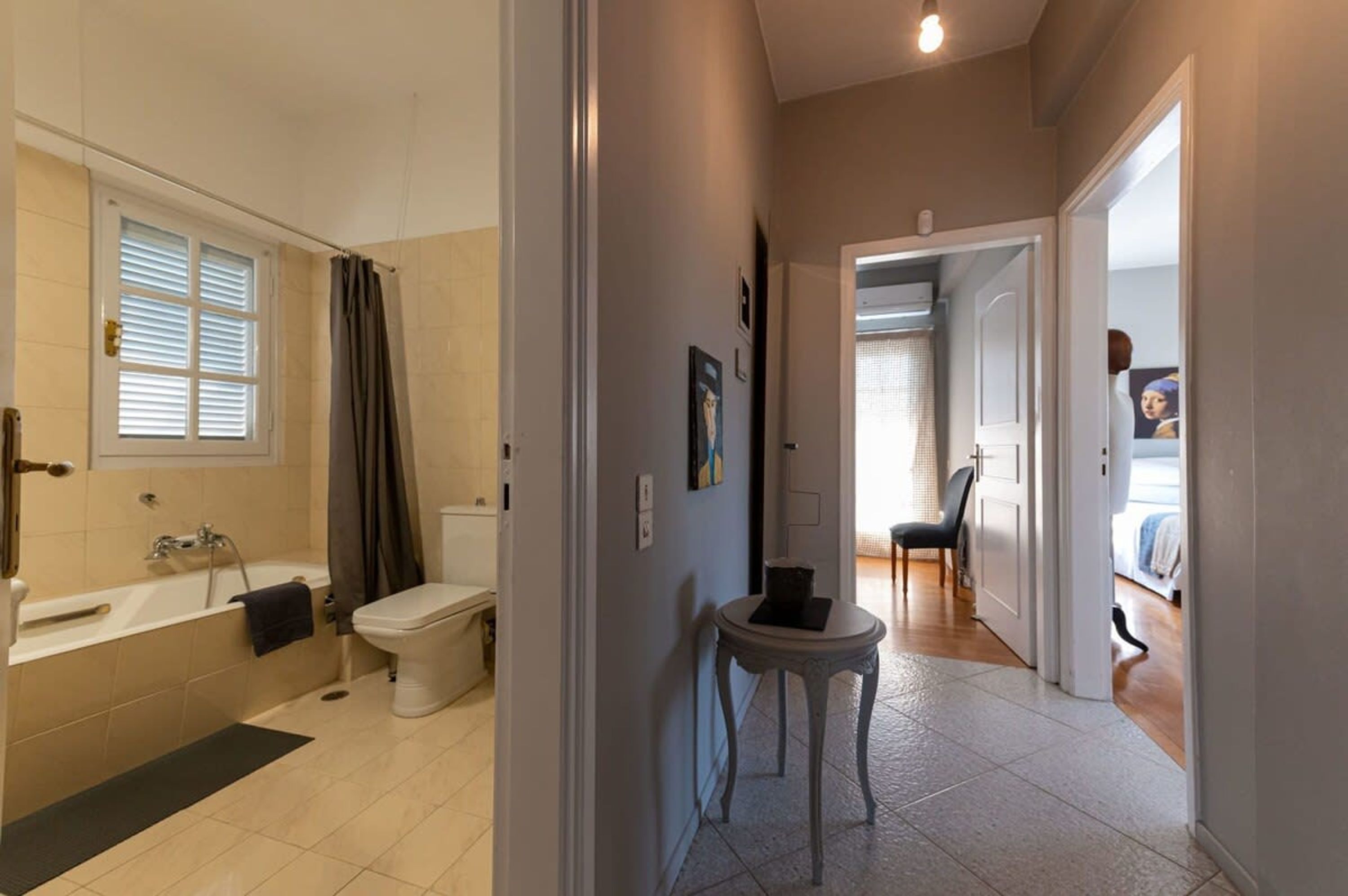 Baño con bañera, inodoro y ventana con persianas. En el pasillo, hay una pequeña mesa con una silla.