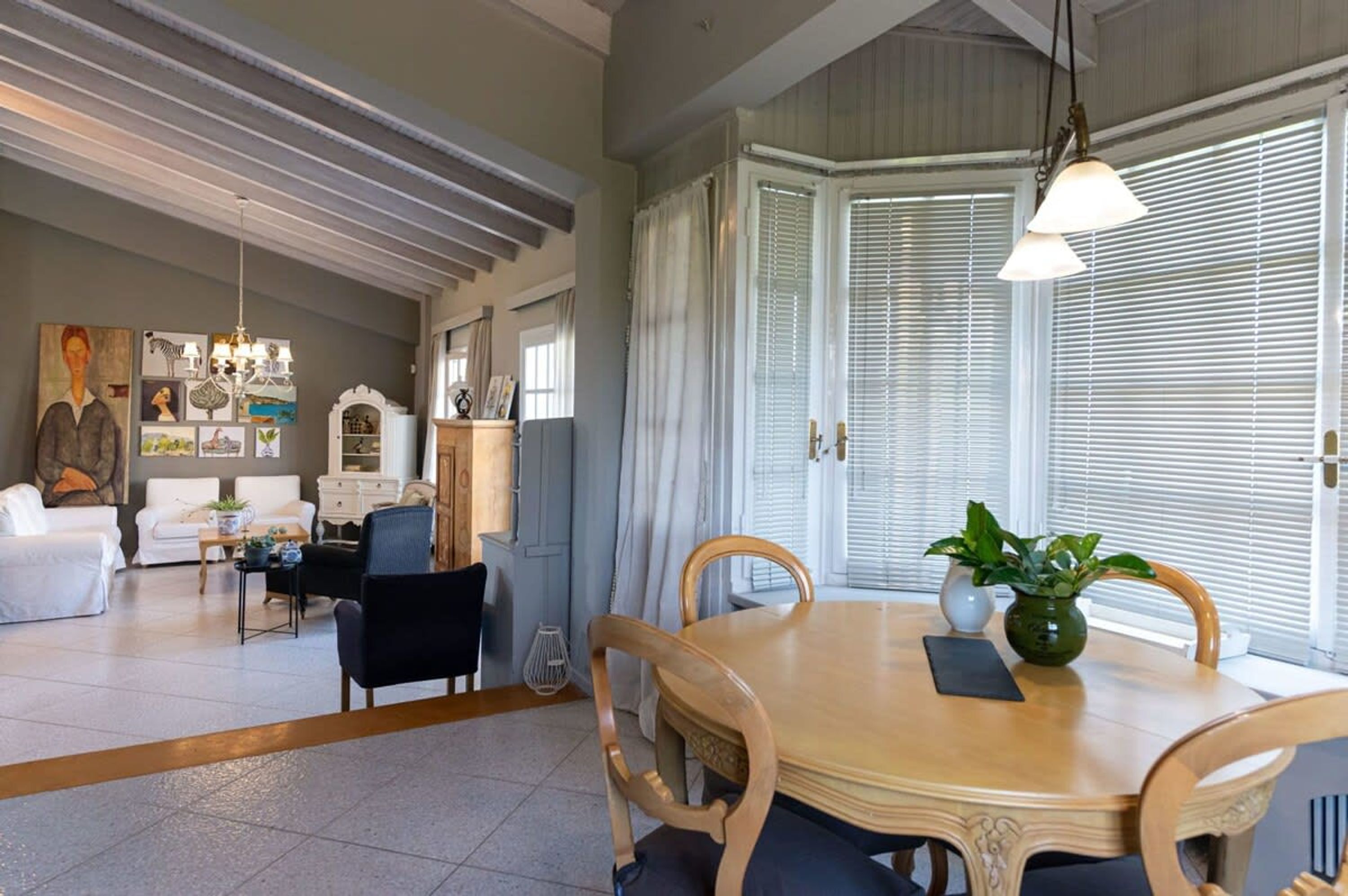 Un'ampia zona giorno con tavolo e sedie da pranzo, illuminata da un lampadario. Una grande finestra con persiane e una porta con tenda.