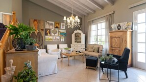 Ein geräumiges Wohnzimmer mit hoher Decke, einem Chandelier und einem großen Fenster. An den Wänden befinden sich mehrere Kunstwerke, ein Holzschrank und verschiedene Sitzgelegenheiten, darunter eine Couch, Stühle und ein Hocker.