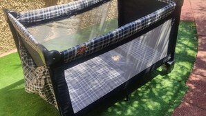 A baby stroller with a mesh seat and a checkered fabric cover, positioned on artificial grass with a stone wall and a paved path in the background. - Selinitsa Stone Studio - Mani Seaview Retreat (West Mani)