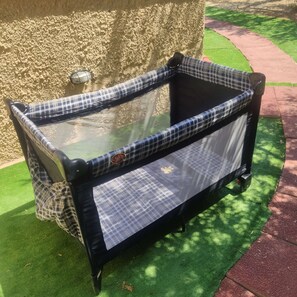 A baby stroller with a mesh seat and a checkered fabric cover, positioned on artificial grass with a stone wall and a paved path in the background.