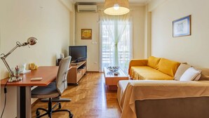 A compact room with a wooden floor, a desk with a computer and lamp, a TV, a sofa, and a hanging light fixture. - Athens Strawberry Hideaway - City Chic Retreat (Athens)