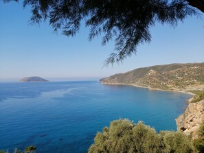Eine Küstenlandschaft mit klarem, blauem Meer, einer kleinen Insel und hügeligem Gelände mit spärlicher Vegetation.