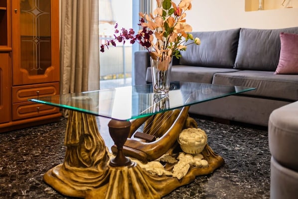 A glass-top table with a decorative base, a vase with flowers, and a sofa in the background.