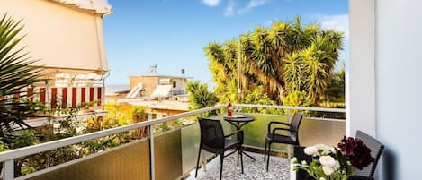 A balcony with a table and chairs, a vase with flowers, and a view of the sea and greenery.