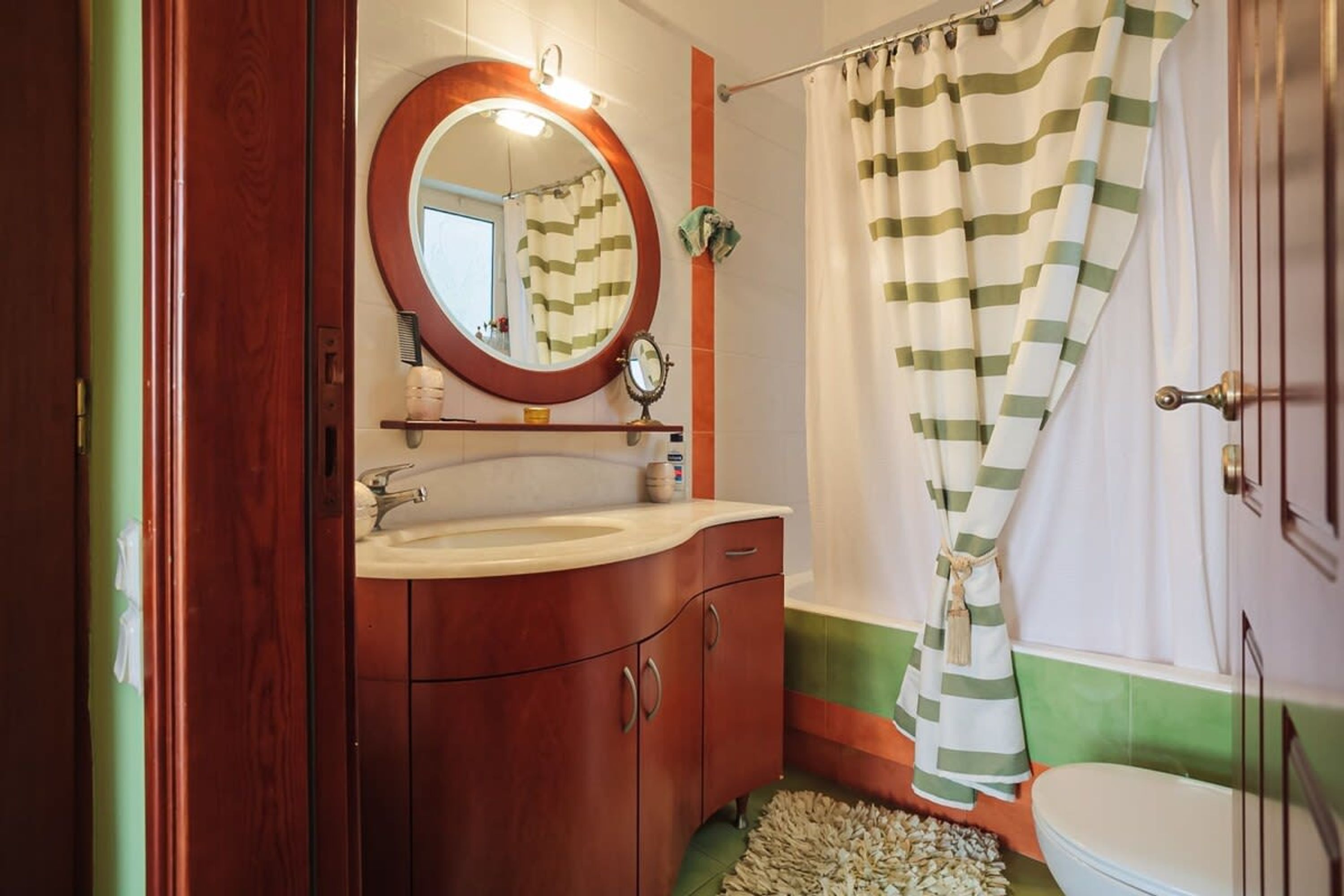 A bathroom with a wooden vanity, a round mirror, a striped shower curtain, and a white toilet.