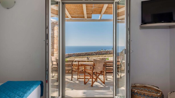 A room with a bed, a television, and a sliding glass door leading to an outdoor seating area with a view of the sea. - Rustic Stone House in the Heart of Folegandros (Folegandros)