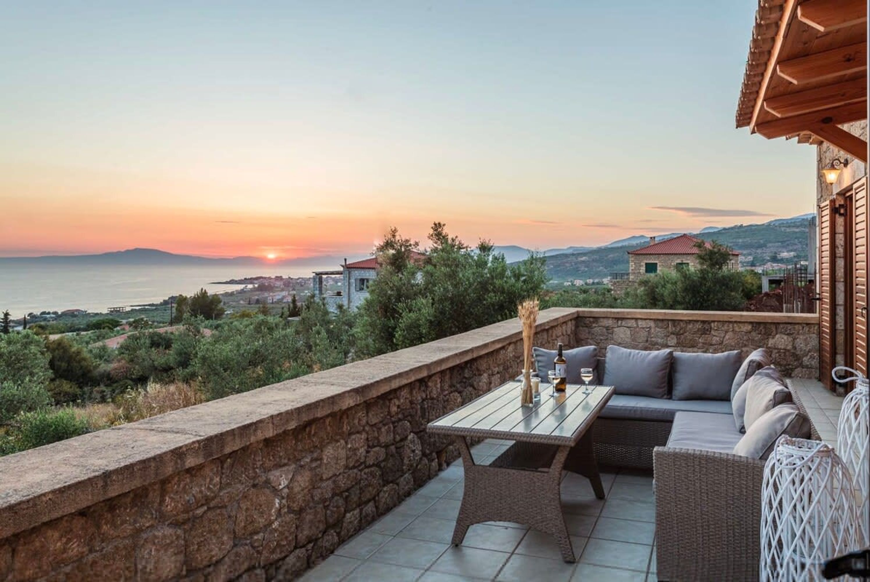 Une terrasse avec un mur en pierre, une table en verre, des meubles en osier et une vue sur le coucher de soleil.