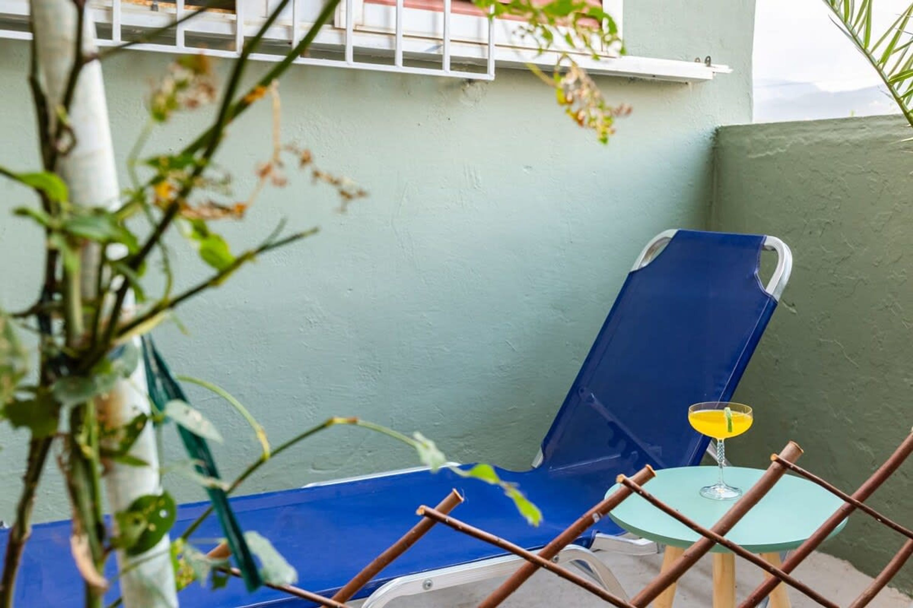 A blue lounge chair with a glass table and a yellow drink, positioned against a light green wall.