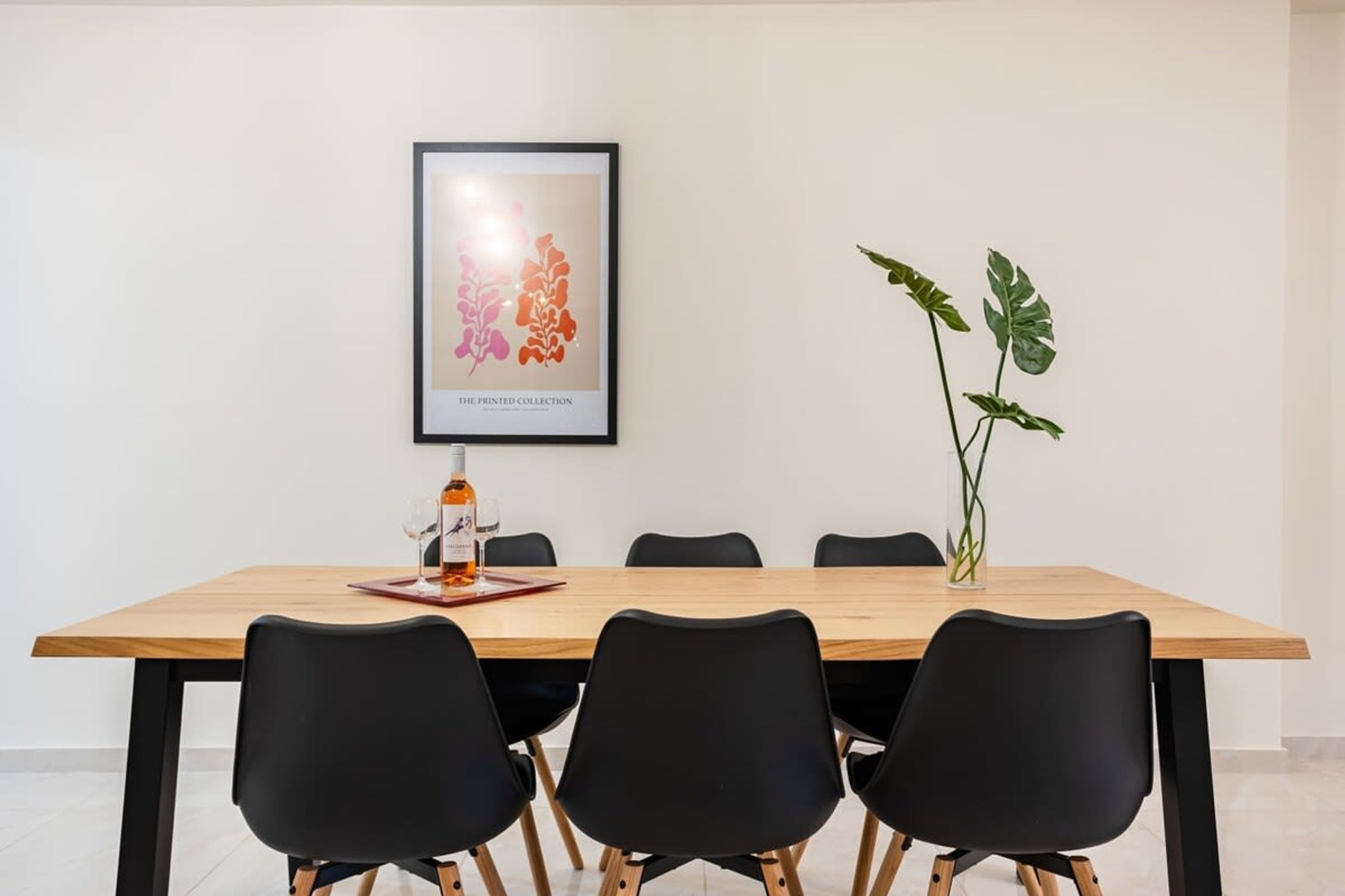 A dining area with a wooden table, black chairs, a framed artwork, and a potted plant.