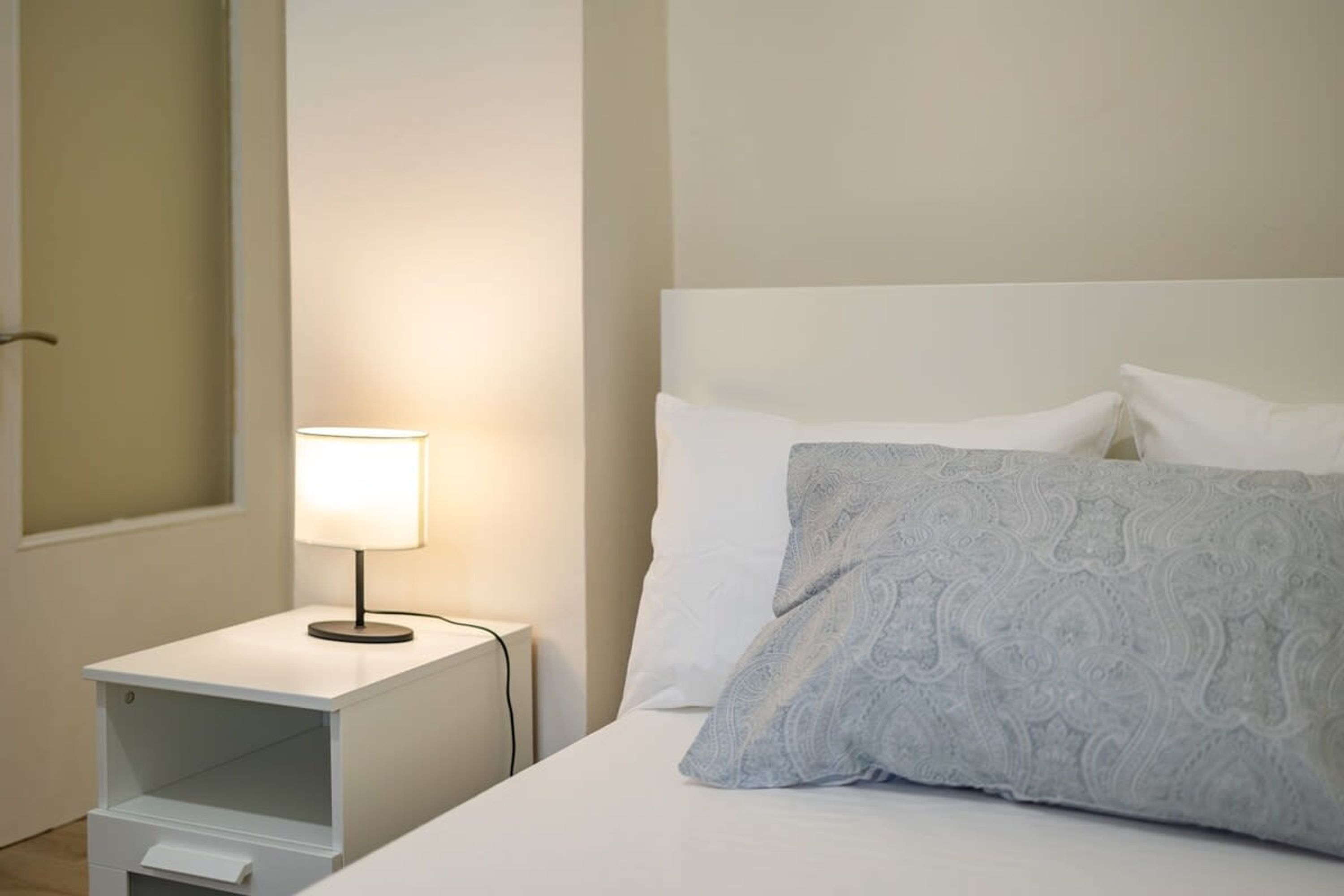 Un lit bien fait, avec du linge de lit blanc et un coussin à motifs, une table de chevet avec une lampe, et un mur sobre.
