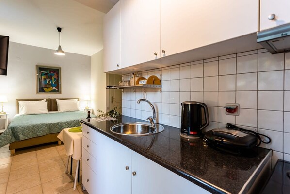 A compact hotel room with a kitchenette, a bed with a green bedspread, a bedside table, and a painting on the wall. - Kitries Summer Getaway - Eden Seaside Suite (West Mani)
