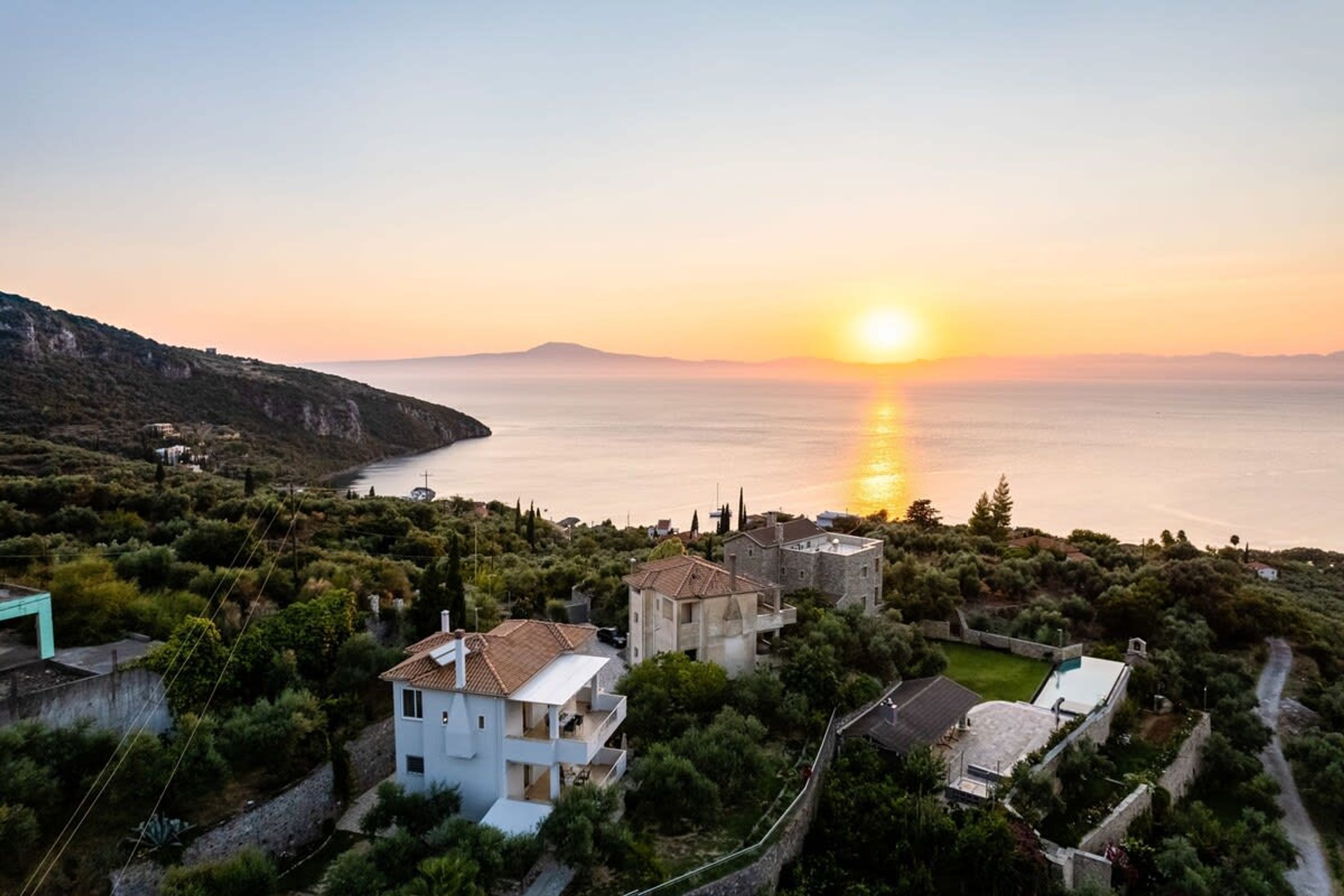 A coastal village with houses surrounded by greenery, a clear view of the sea, and a sunset in the background.