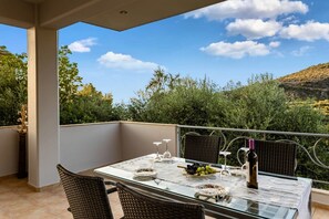 A balcony with a glass table set for dining, wicker chairs, and a view of greenery and hills.