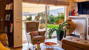 A living room with a sofa, armchair, and coffee table. There is a TV mounted on the wall and a bookshelf with books. - Sunset Paradise - Verga s Ultimate Seaview Getaway (Kalamata)