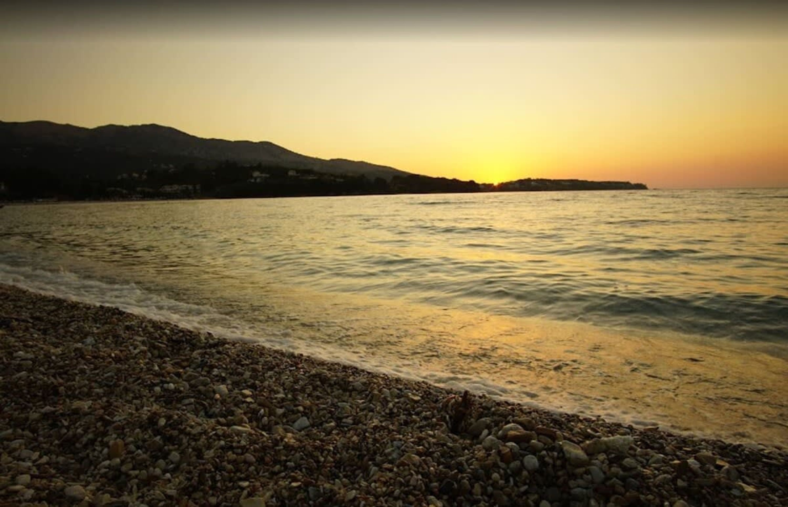 Une plage de galets, avec un coucher de soleil sur l’eau et des collines lointaines.