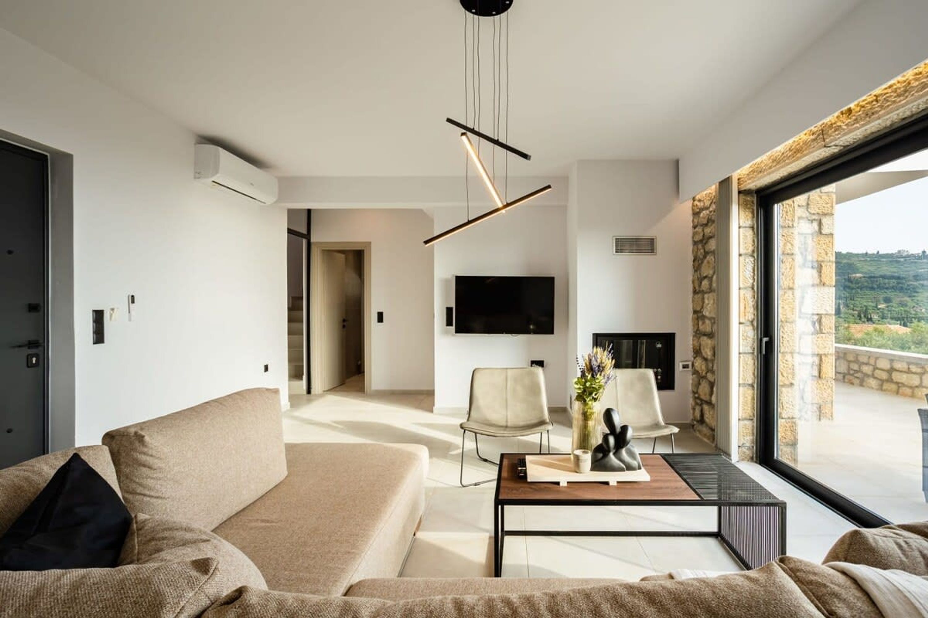 A modern living room with a sofa, chairs, a coffee table, and a TV. There is a stone wall and a large window with a view.