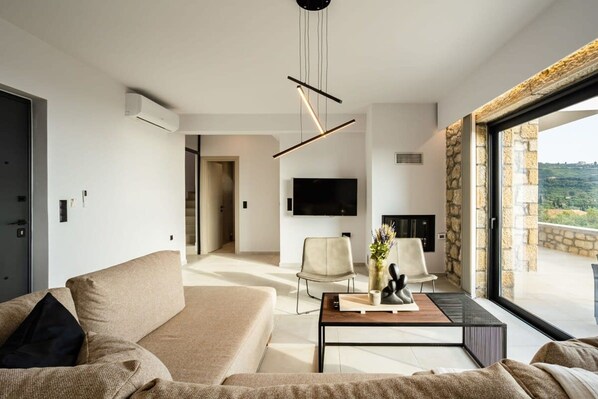 A modern living room with a sofa, chairs, a coffee table, and a TV. There is a stone wall and a large window with a view. - Mani Stone Villa A Paradise of Panoramic Views (West Mani)
