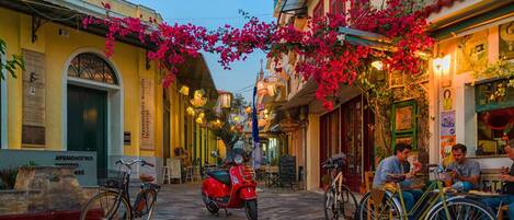 A narrow street with buildings on either side, a red scooter parked in the middle, and people sitting at outdoor tables.